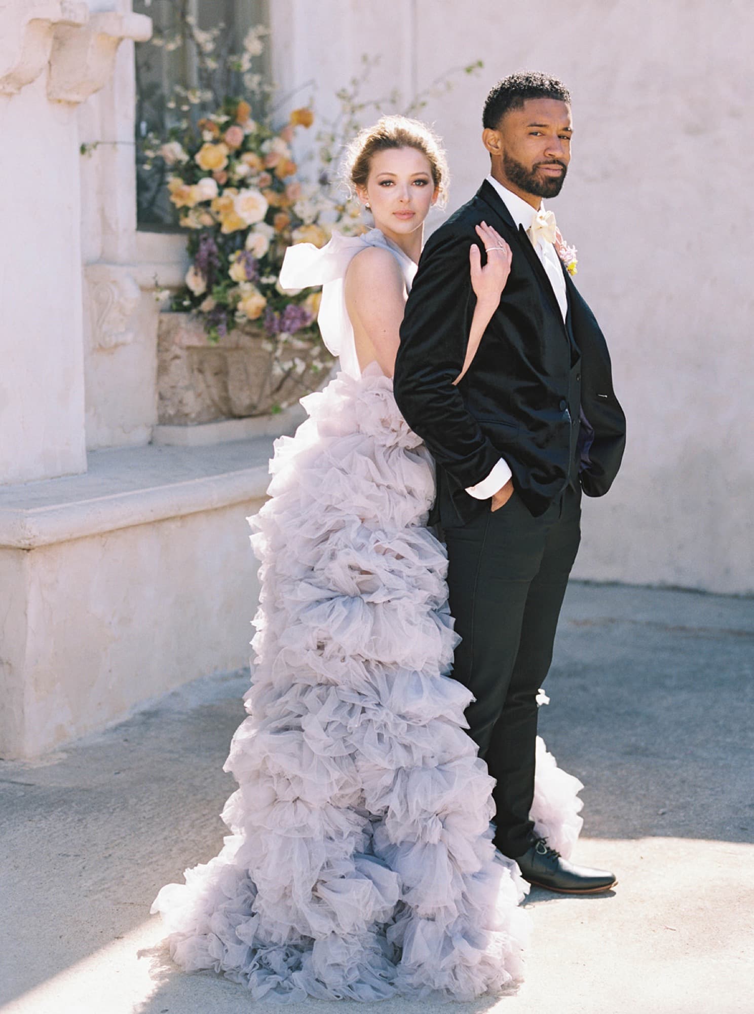 Abyon Ausar in a black velvet tuxedo standing back to back with a bride in a ruffled lilac gown