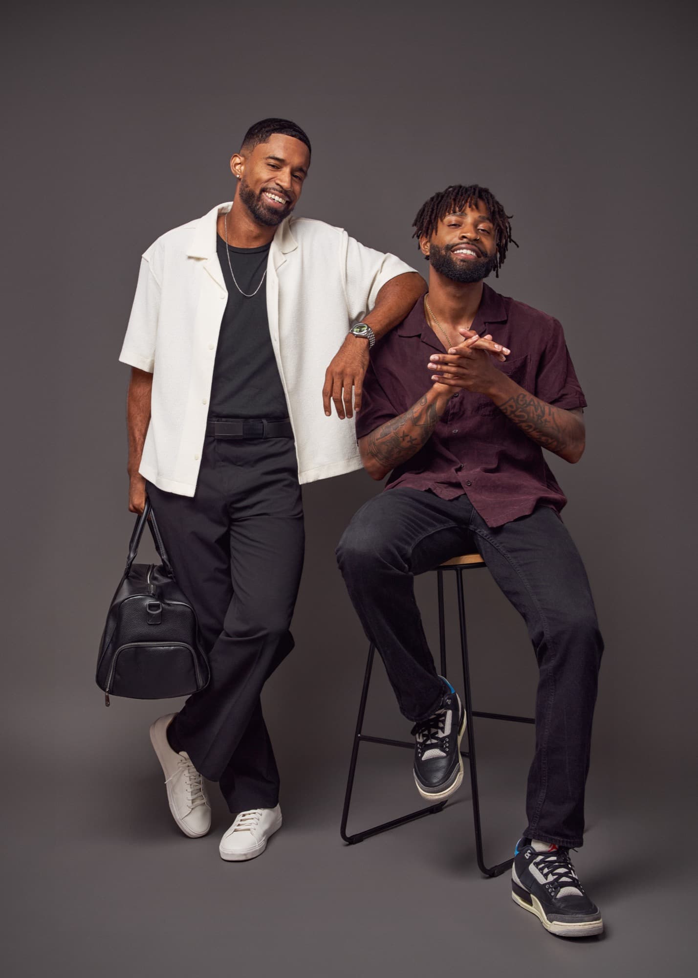 Abyon Ausar holding a leather duffel bag beside another model on a stool