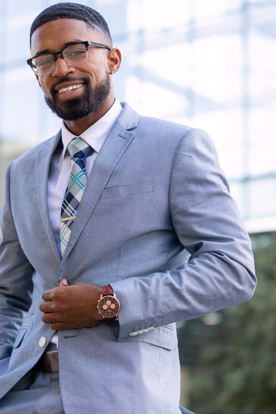 Abyon Ausar buttoning a light grey three-piece suit outdoors, watch on his wrist