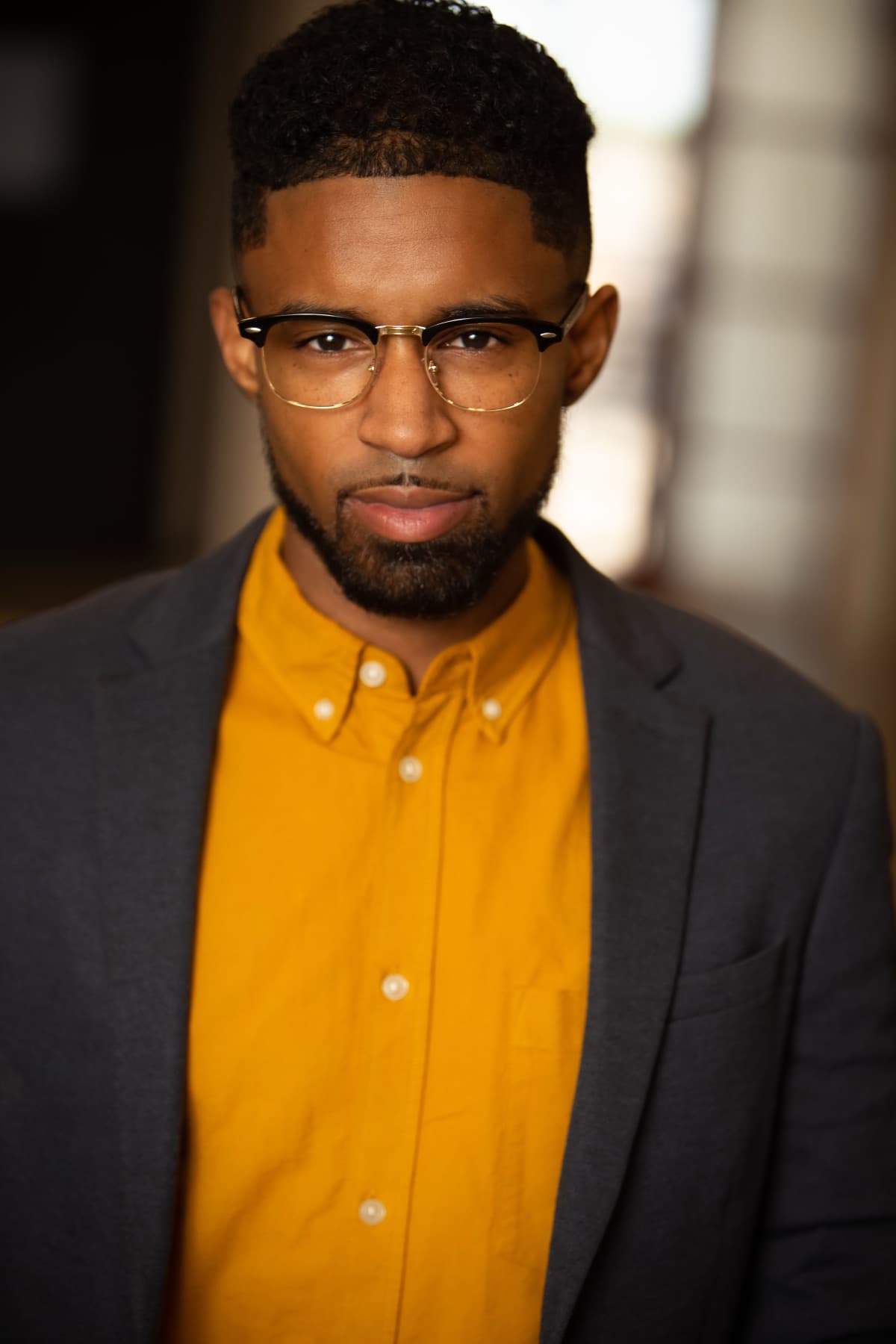 Headshot of Abyon Ausar in a grey blazer, mustard shirt and glasses