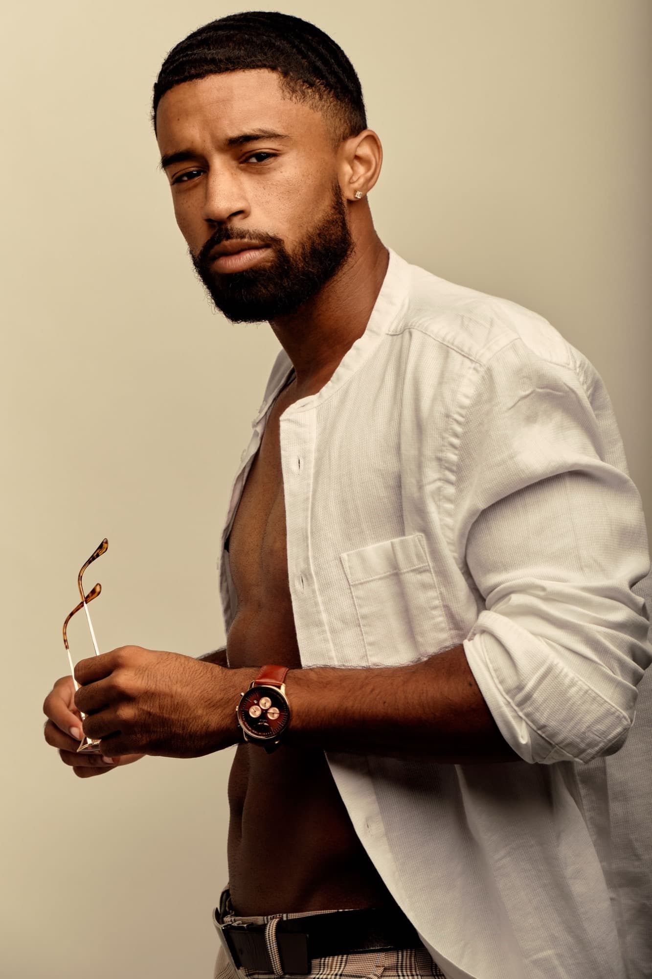 Abyon Ausar in an open white shirt against a cream backdrop, holding sunglasses