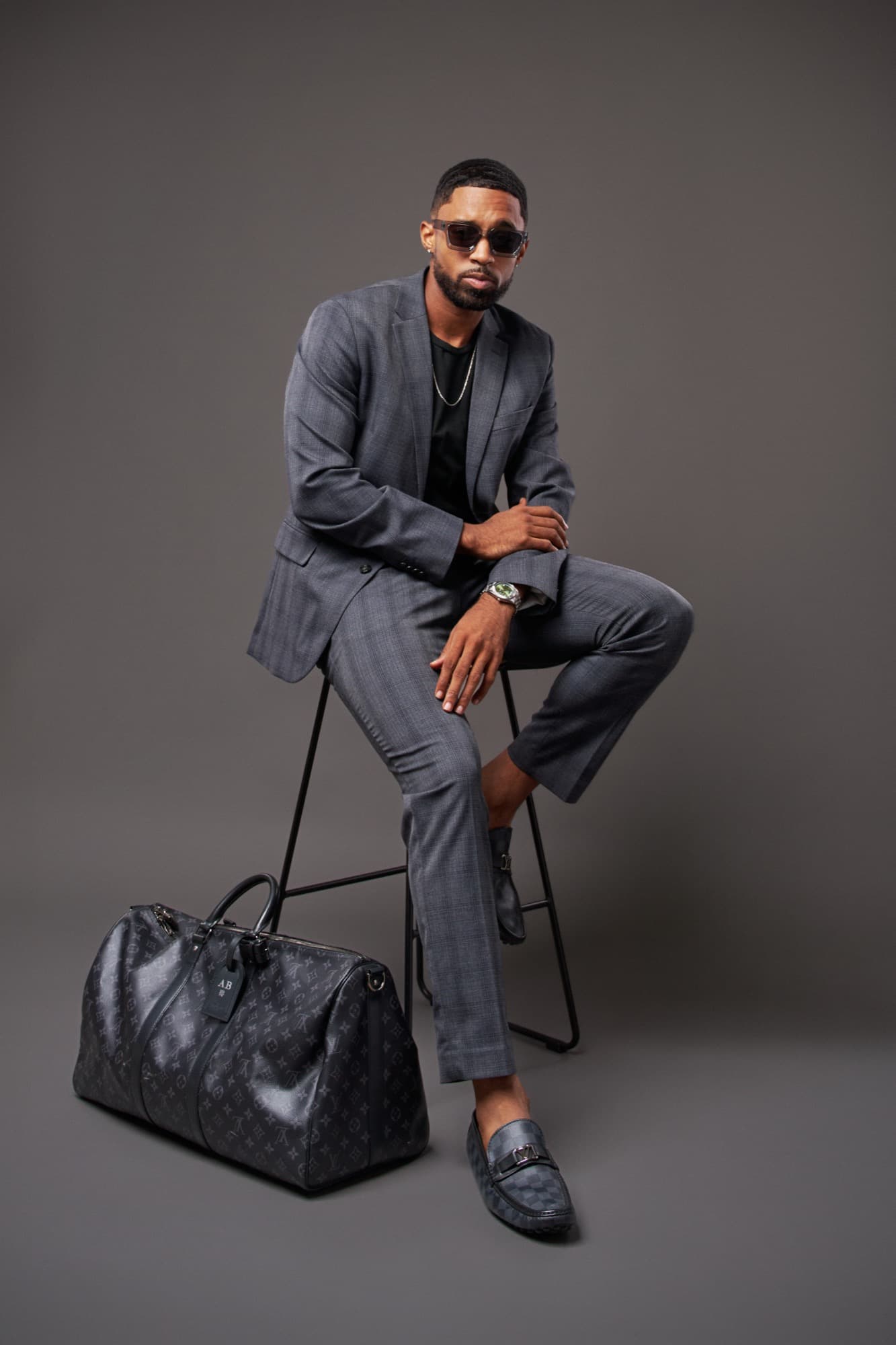 Abyon Ausar seated in a grey checked suit and sunglasses beside a leather holdall