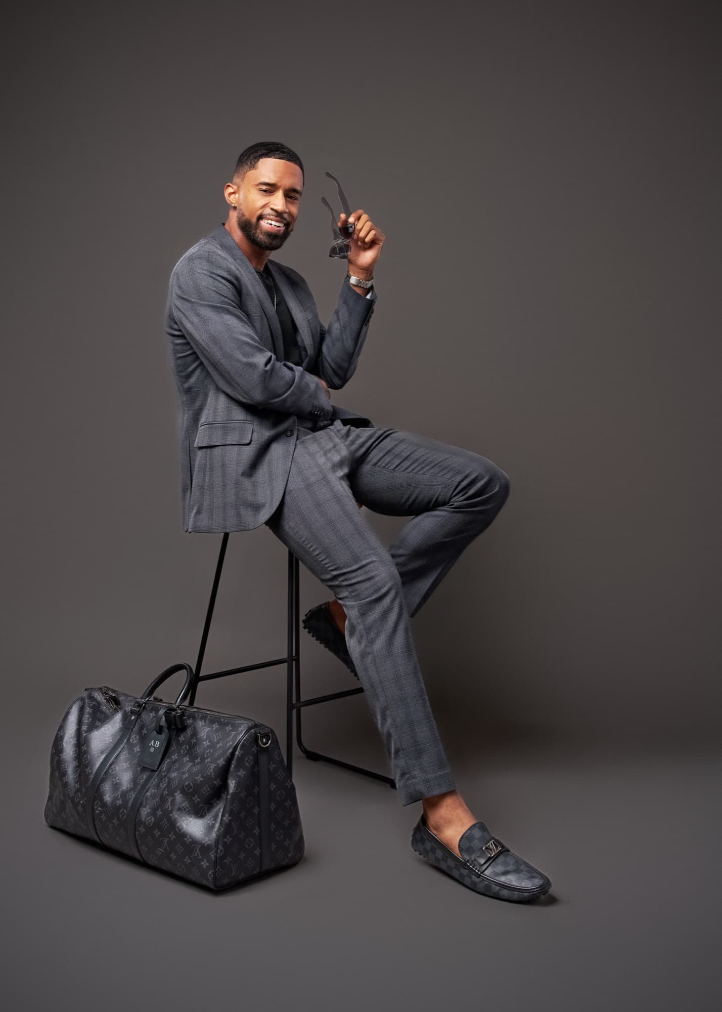Abyon Ausar seated in a grey checked suit, smiling, holding his sunglasses
